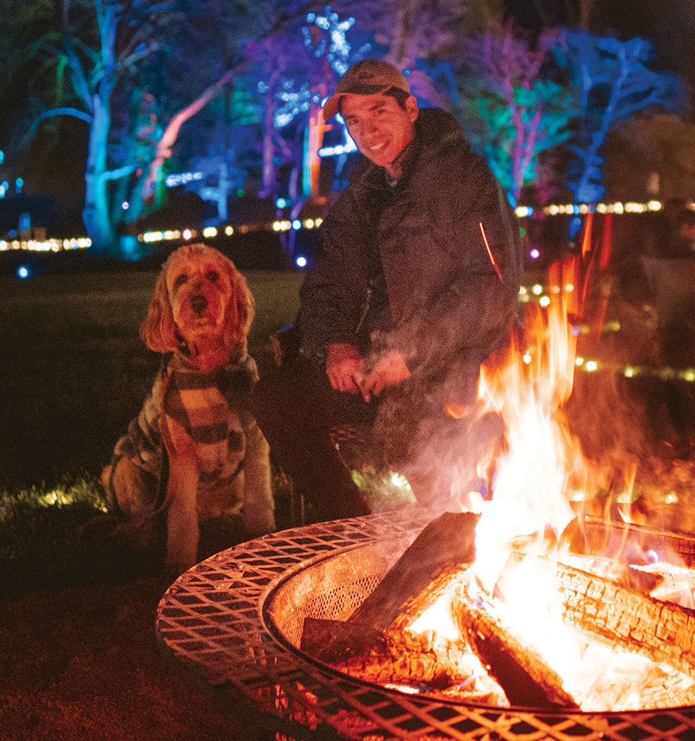 A man grills by a roaring fire pit at night while a golden retriever and another person watch city lights behind them.