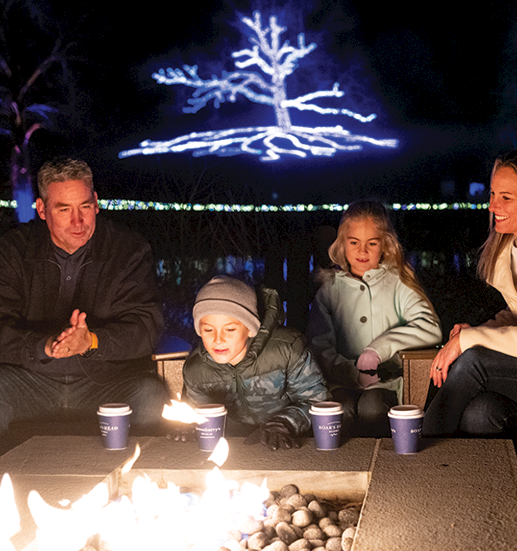 A family sits by a glowing outdoor fire at night, sipping drinks with colorful lights and an illuminated tree above.
