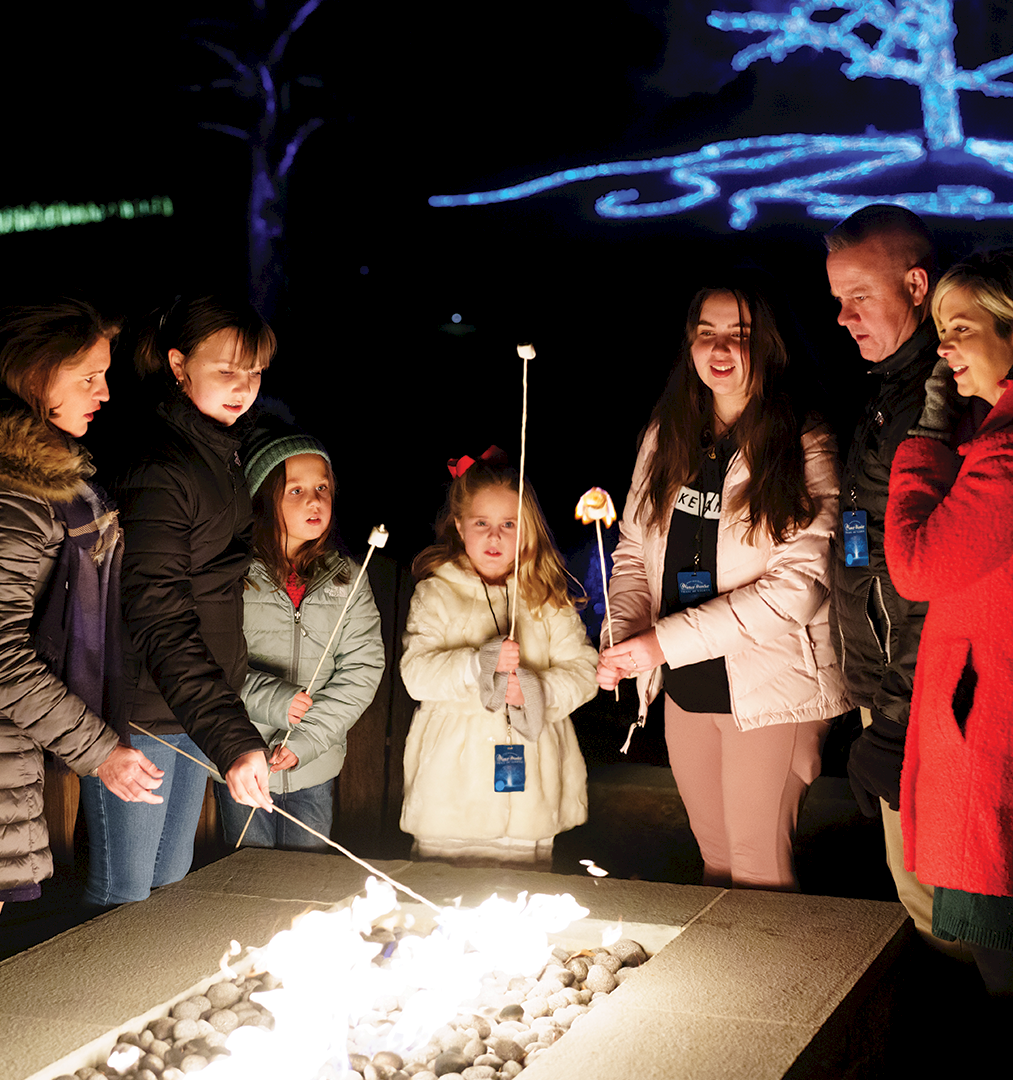 A group of people roast marshmallows around a glowing campfire table at night, with colorful light projections overhead.