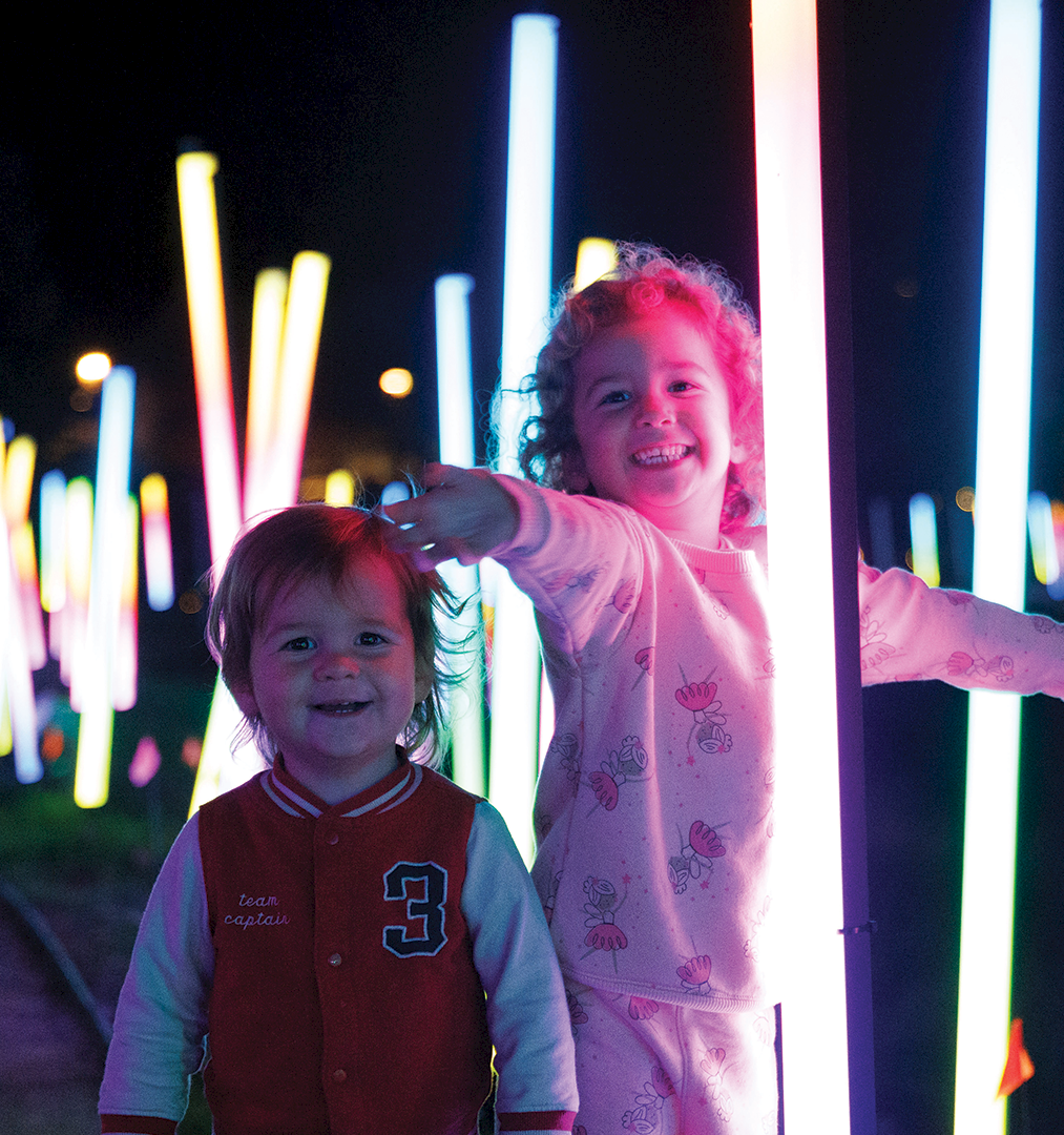 Two kids smile in front of colorful glowing light sticks at night, with bright neon bars stretching across the background.