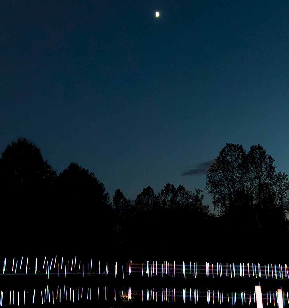 Night sky with a bright moon above dark trees and a river, with colorful light reflections along the water in the foreground.