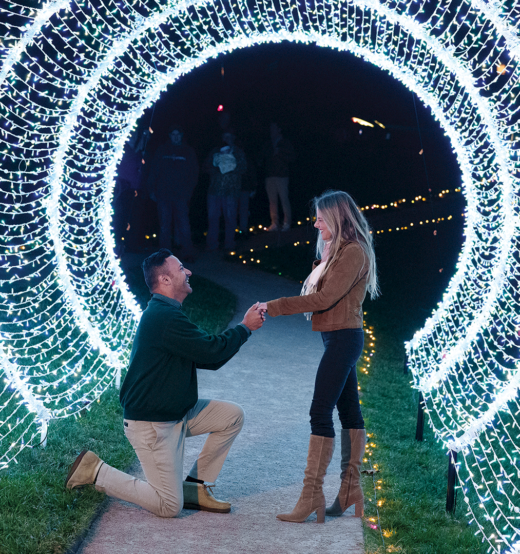 A man kneels proposing to a woman under glowing light tunnels at night during a wedding-style celebration.