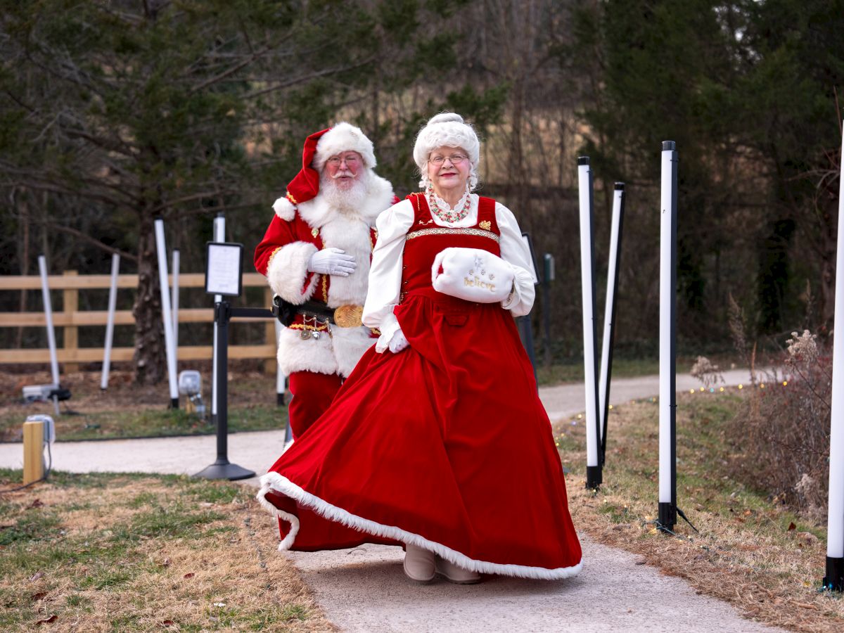 A man dressed as Santa and a woman in festive red attire stand together outdoors by event lights and speakers, smiling.
