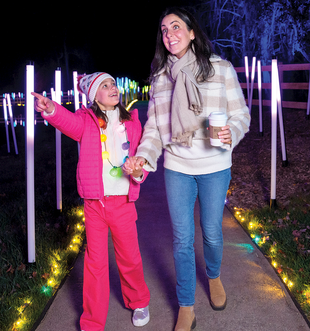 Two people in winter clothes stroll at night by colorful light tubes, with the little girl pointing and the older woman holding a coffee.