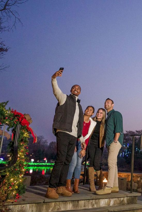 Friends take a selfie by a giant glowing Christmas tree at night, surrounded by holiday lights and decorations.