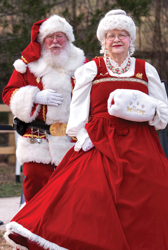 Santa and Mrs. Claus in red outfits stand outdoors on a path beside event light poles, smiling.