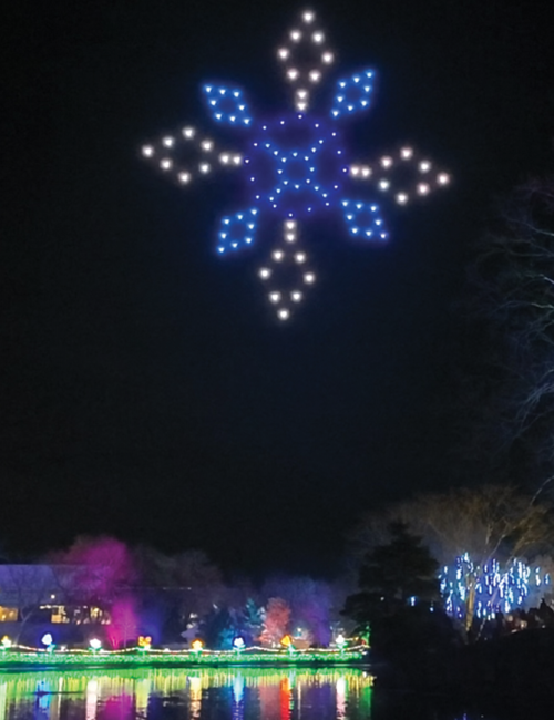 A drone light display forms a glowing star above a colorful reflection in the water.