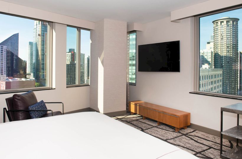 A modern bedroom with a large bed, TV on the wall, and wide windows showing a tall city skyline.