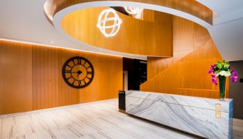Modern hotel lobby with a marble reception desk, warm wood walls, wall clock, hanging globe lights, and a flower vase.