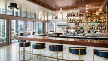 A modern bar interior with a stocked liquor shelf, glowing hanging lights, and a marble counter with blue-and-gold stools.