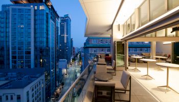 Modern office rooftop balcony with cozy seating, looking over a city street lined with skyscrapers at dusk.