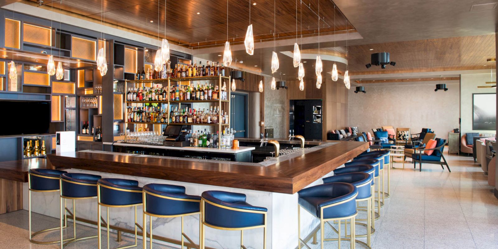 Modern hotel bar interior with a wooden counter, glowing bottles on shelves, hanging lights, stools, and lounge seating beyond.