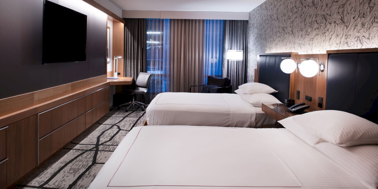 A modern hotel room with twin beds, wood paneling, curtains, and bedside lamps by the headboard.