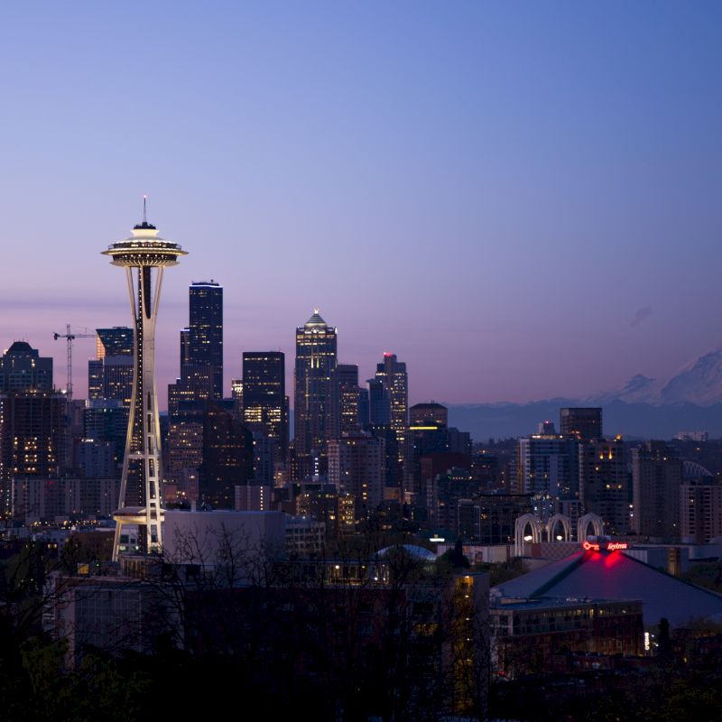 Seattle&rsquo;s skyline at dusk with the Space Needle lit up, surrounding skyscrapers, and distant mountains under a purple sky.