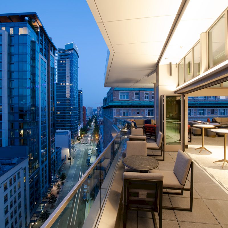 Rooftop terrace with lounge chairs overlooks a city skyline of modern office buildings at dusk.