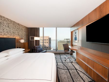 Modern hotel room with a large bed, wood-paneled walls, chair and desk, big TV, and a city view through large windows.