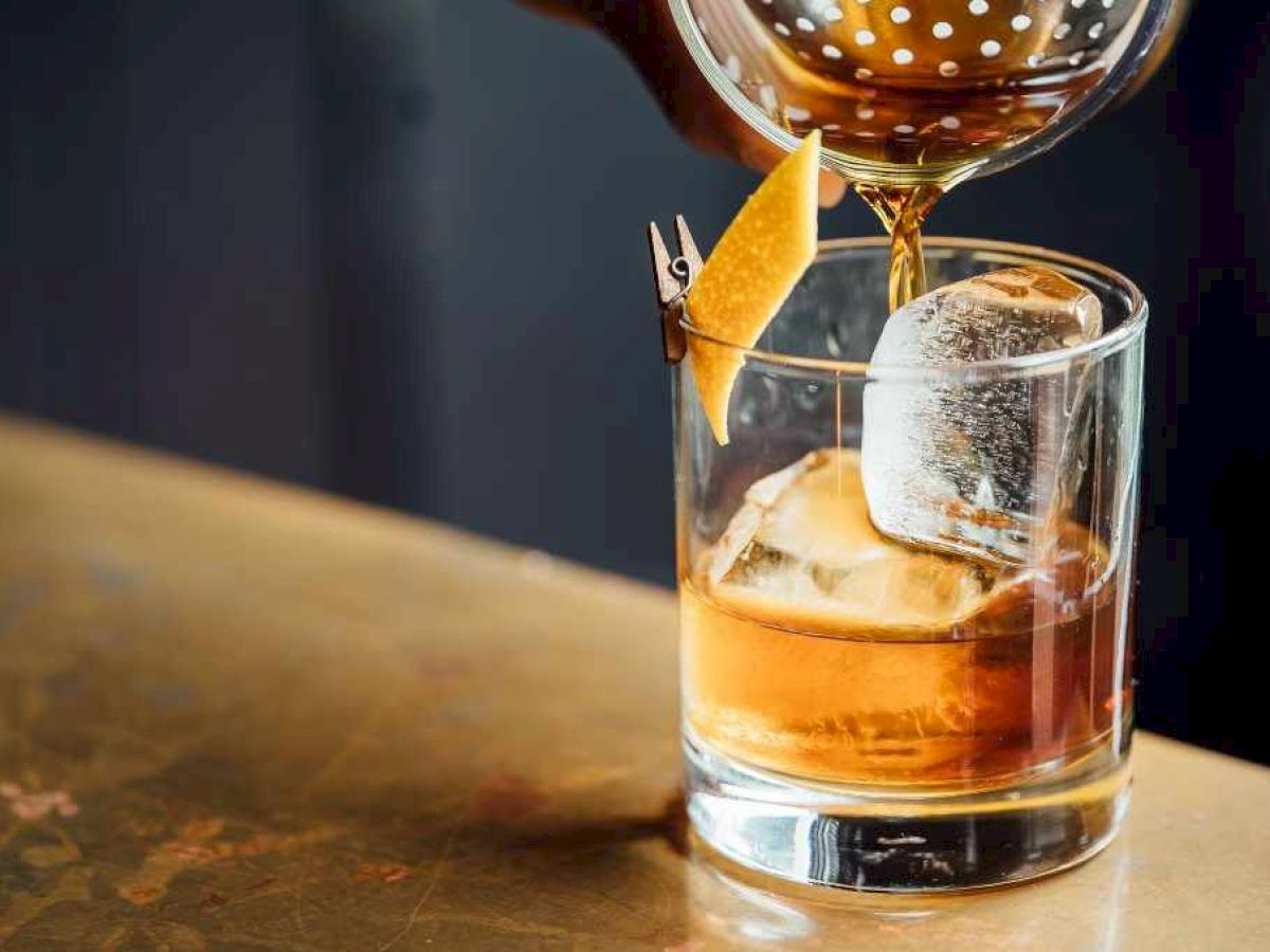 Amber liquor is being poured into a rocks glass with ice and a citrus wedge garnish.