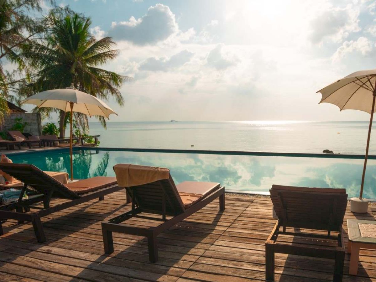 A tropical oceanfront resort deck with a pool, lounge chairs, and umbrellas beside the calm sea under a bright sky.