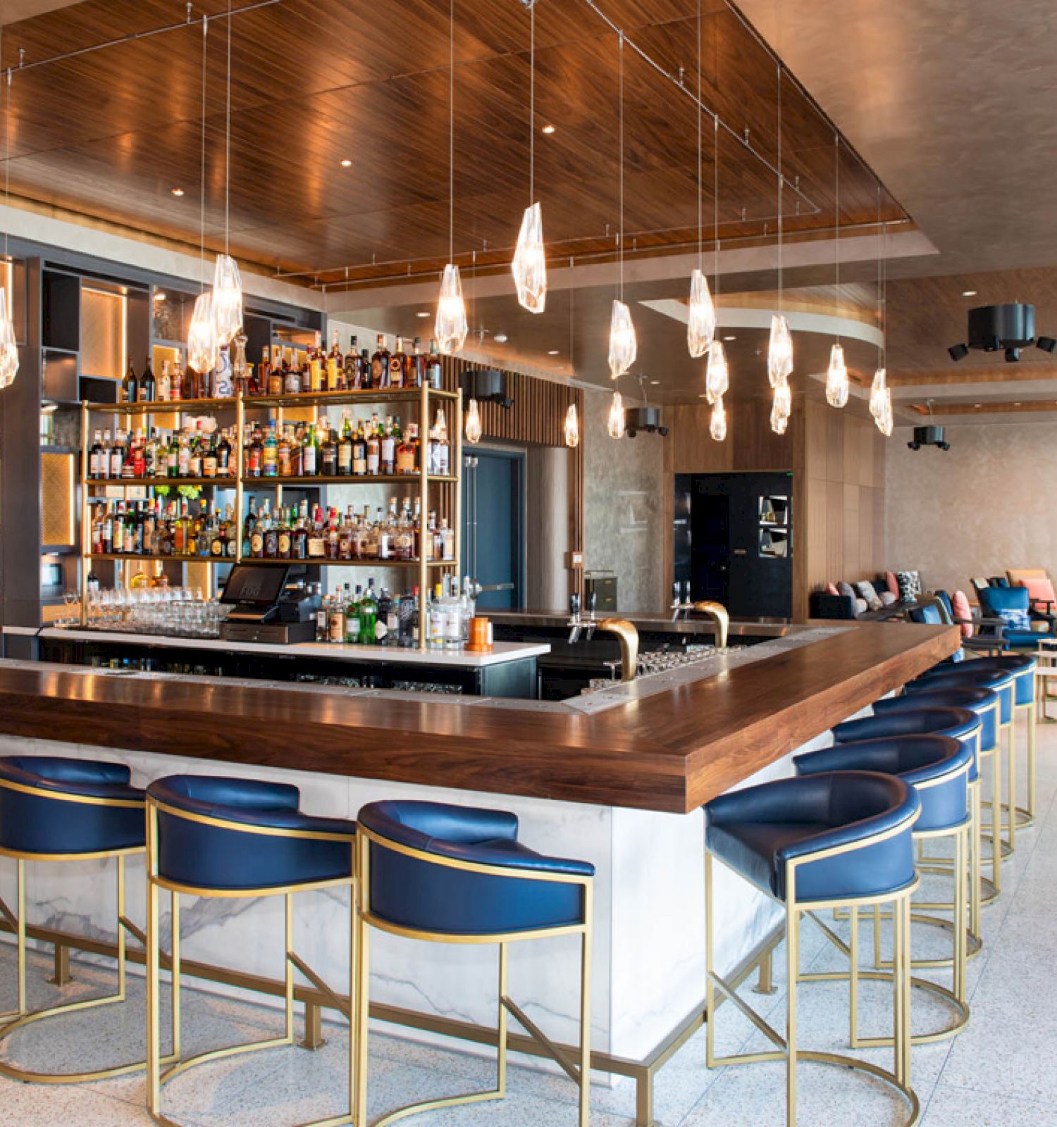 A modern hotel bar with a stocked bottle wall, marble counter, blue stools, and glowing pendant lights, plus a lounge seating area.