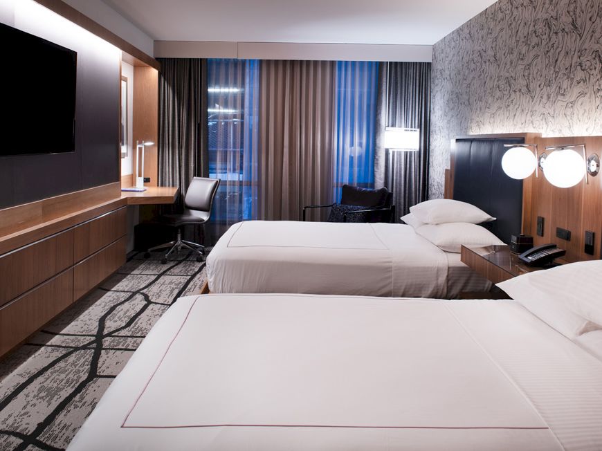 A modern hotel room with two beds, a wall-mounted TV, wood paneling, and floor-to-ceiling curtains by a window, neatly staged.