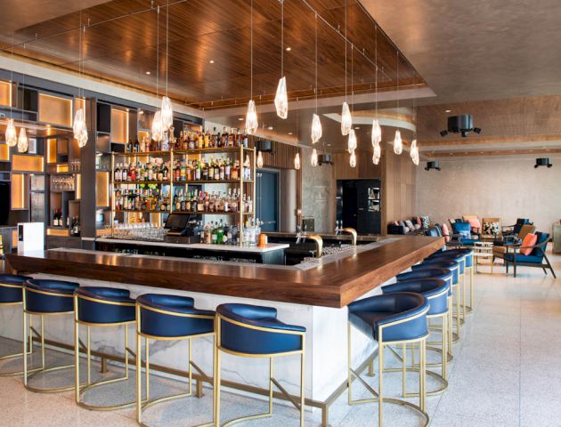 A modern hotel bar with a stocked bottle wall, sleek counter, blue stools, hanging lights, and a lounge seating area in view.