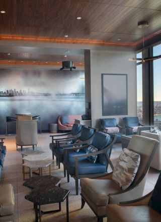 A modern hotel lounge with plush chairs, floor lamps, huge window views of a river and port, and a cityscape mural.