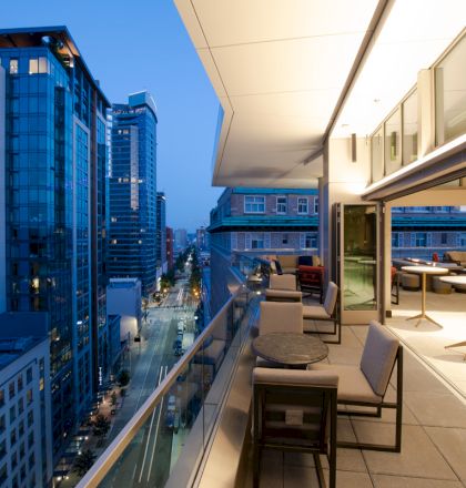 Balcony lounge overlooks a downtown street at dusk with tall buildings and city lights.