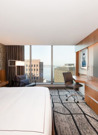 A modern hotel bedroom with a king bed, chair, desk area, TV wall, and large window overlooking city buildings.