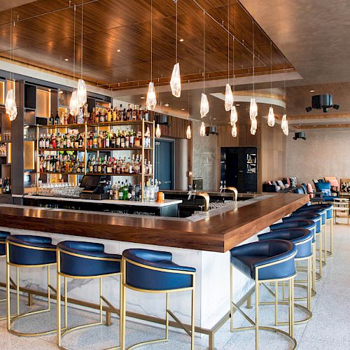 A modern lounge bar interior with a wood counter, stocked bottles, pendant lights, and blue high stools and lounge seating.