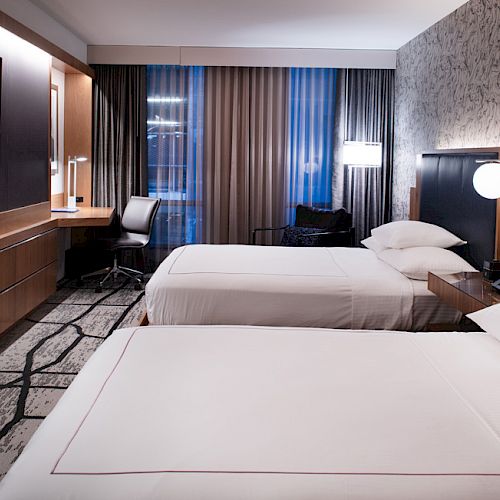 A modern hotel room with two neatly made beds, dark wood panels, and bedside lamps beside a large window.
