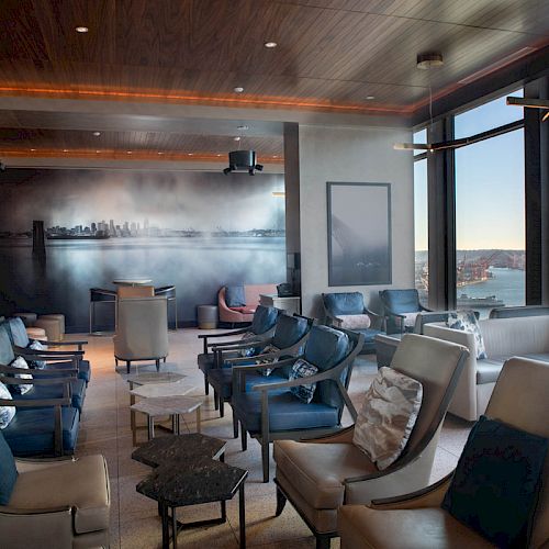 Modern lounge with blue upholstered chairs and tables, large windows overlooking a harbor at dusk.