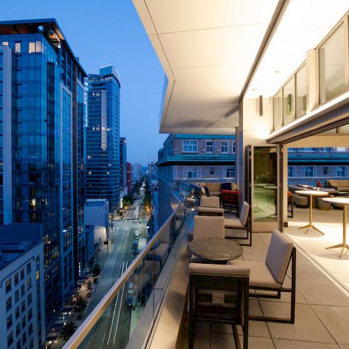 Rooftop terrace with lounge seating overlooks tall modern office buildings down a city street at dusk.