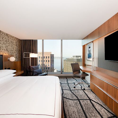 Modern hotel room: large bed, wood cabinets and desk chair, wall TV, and big windows showing a city view.