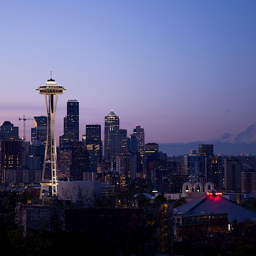 A twilight city skyline featuring Seattle&rsquo;s Space Needle, glowing buildings, and distant mountains under a purple-blue sky.