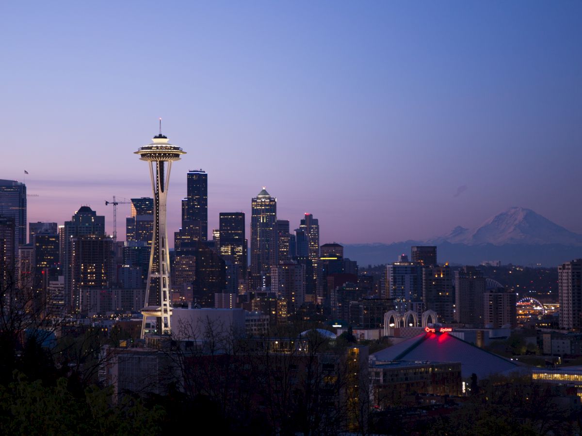 Seattle skyline at dusk with the Space Needle lit up, city buildings below, and distant mountains under a purple sky.