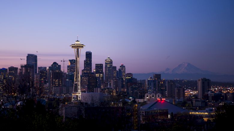 Seattle skyline at dusk with the Space Needle lit up, city buildings below, and distant mountains under a purple sky.