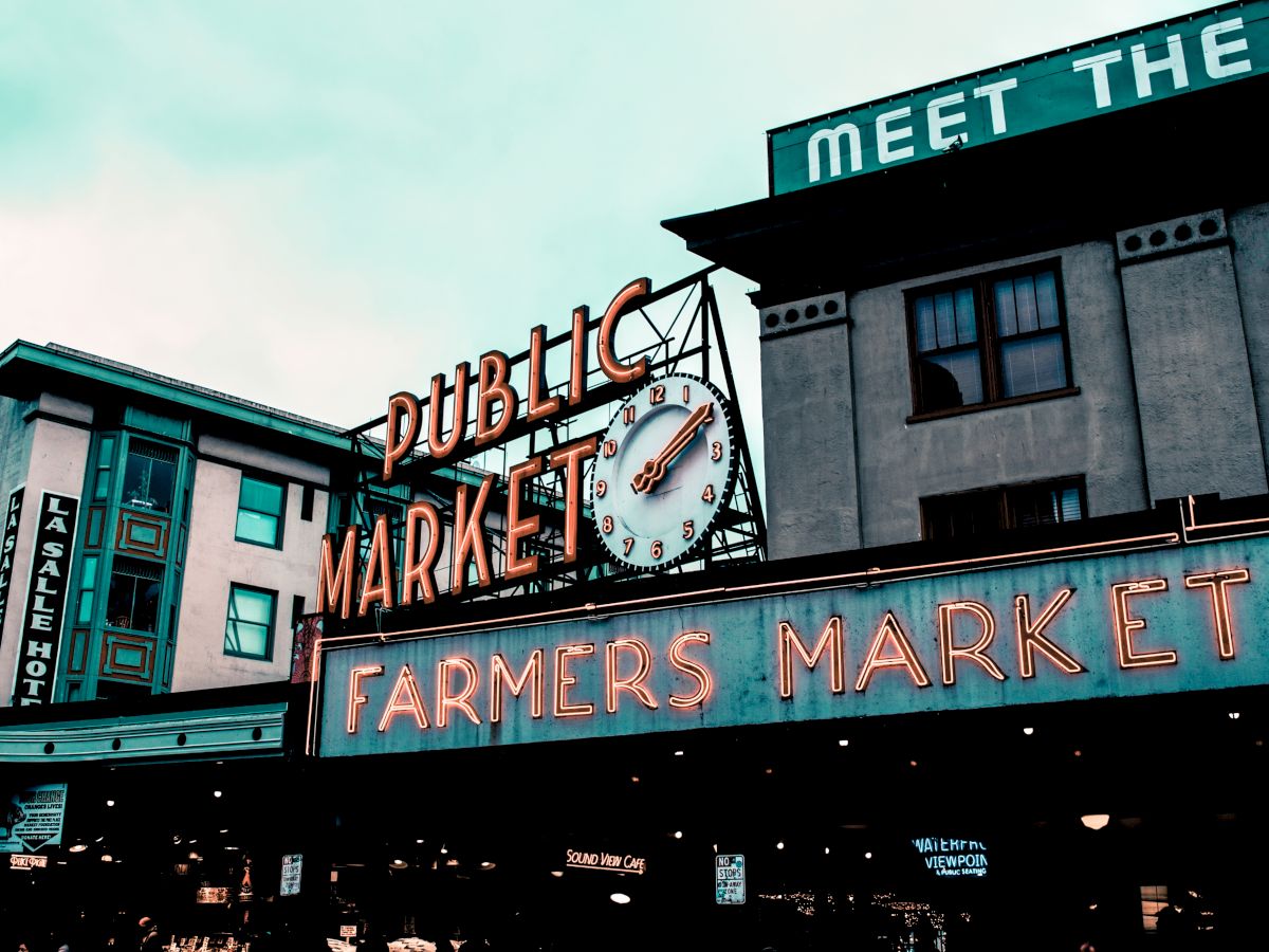 Neon &ldquo;PUBLIC MARKET&rdquo; and &ldquo;FARMERS MARKET&rdquo; signage with &ldquo;MEET THE&rdquo; and a clock on brick buildings.