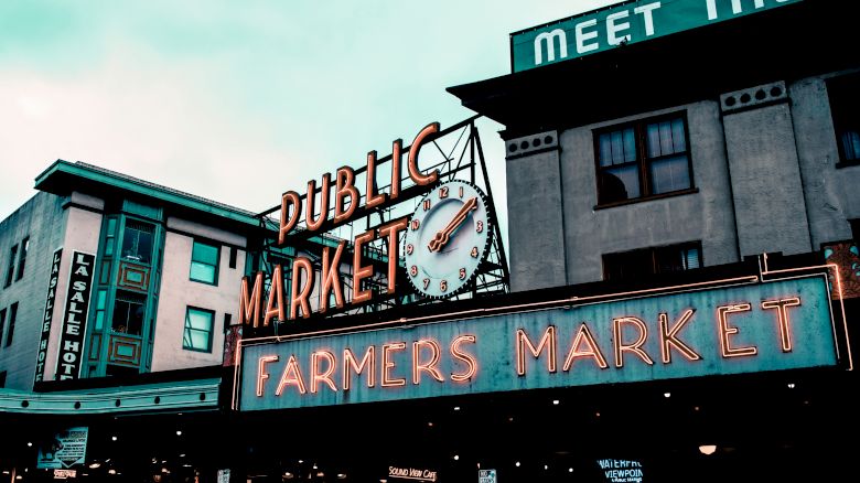 Neon &ldquo;PUBLIC MARKET&rdquo; and &ldquo;FARMERS MARKET&rdquo; signage with &ldquo;MEET THE&rdquo; and a clock on brick buildings.