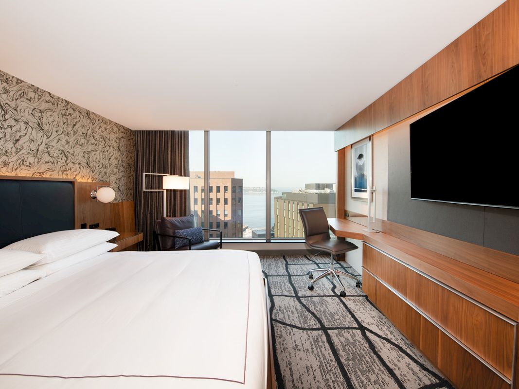 Modern hotel room with a large bed, chair/desk area, wall-mounted TVs, and city-view windows, with wood paneling and carpet.
