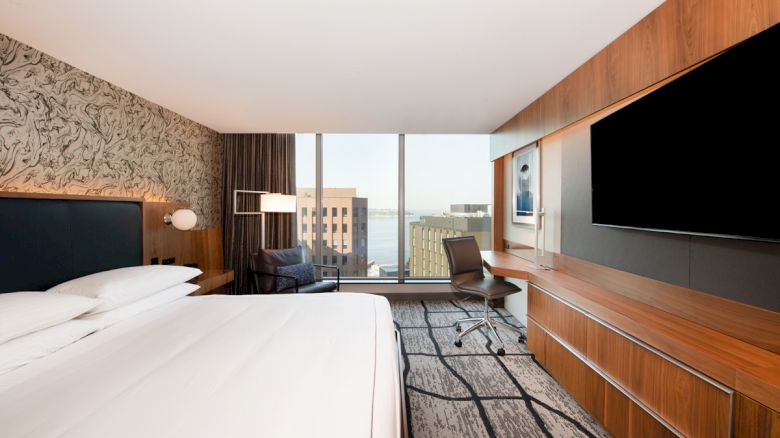 Modern hotel room with a large bed, chair/desk area, wall-mounted TVs, and city-view windows, with wood paneling and carpet.