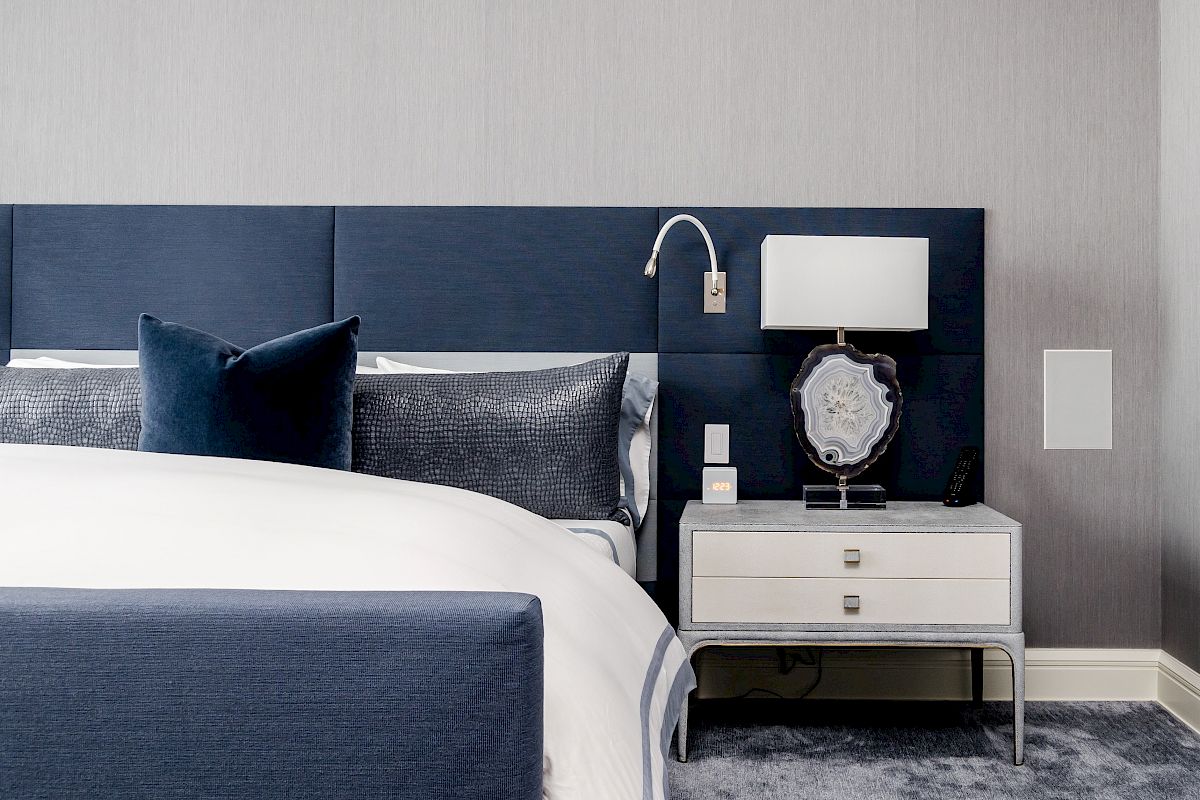 A modern hotel bedroom with a blue bed, pillows, bedside table, lamp, and wall outlets.