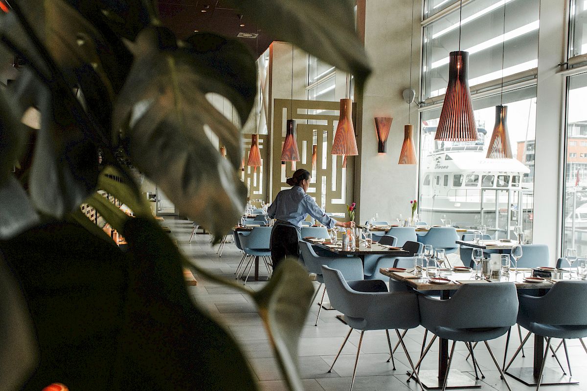 Restaurant dining area with blue chairs, orange pendant lights, a waiter, and a window showing a white boat outside.