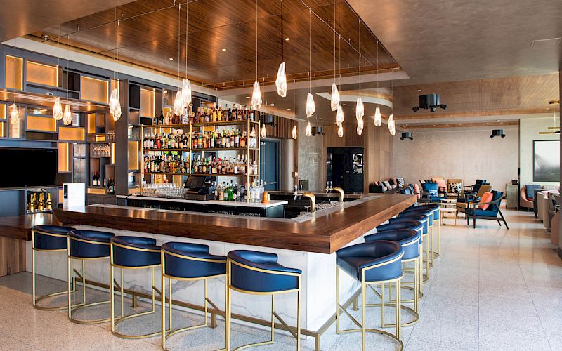 Modern bar interior with backlit shelves of bottles, hanging pendant lights, high stools, and lounge seating in a contemporary room.