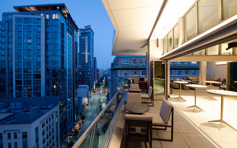 Modern apartment balcony with outdoor seating overlooking downtown buildings under a twilight-blue sky.