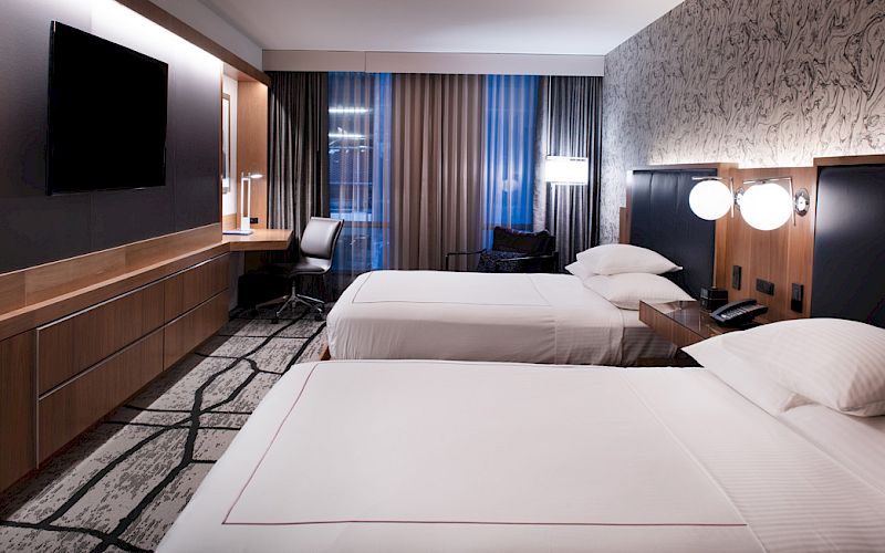 A modern hotel room with two beds, a wall-mounted TV, patterned carpet, curtains, and bedside lamps, lit in warm light.