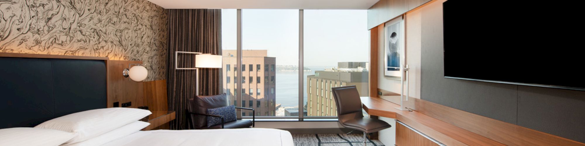 Modern hotel room with a king bed, seating, desk chair, wall TV, wood cabinetry, and large windows overlooking buildings.