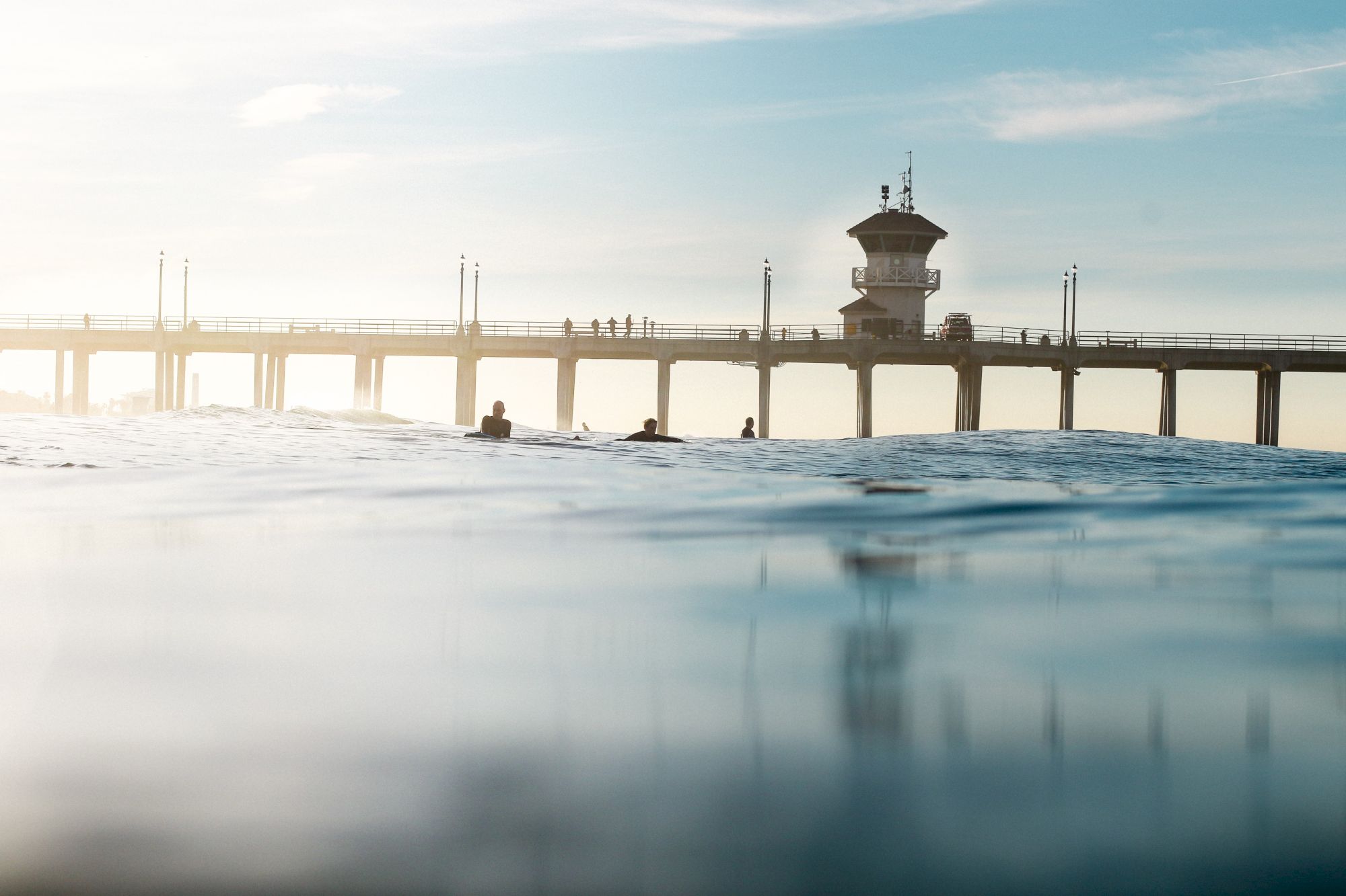 A calm ocean scene with people swimming in waves beneath a seaside pier and a tall lookout tower at sunset.